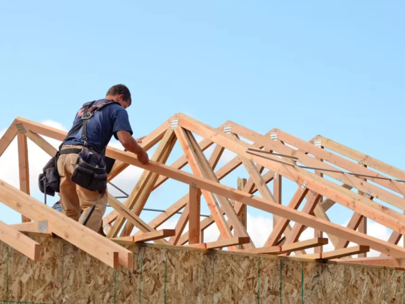 A construction worker installing wooden pole barn trusses on the roof of a new home under a clear blue sky.